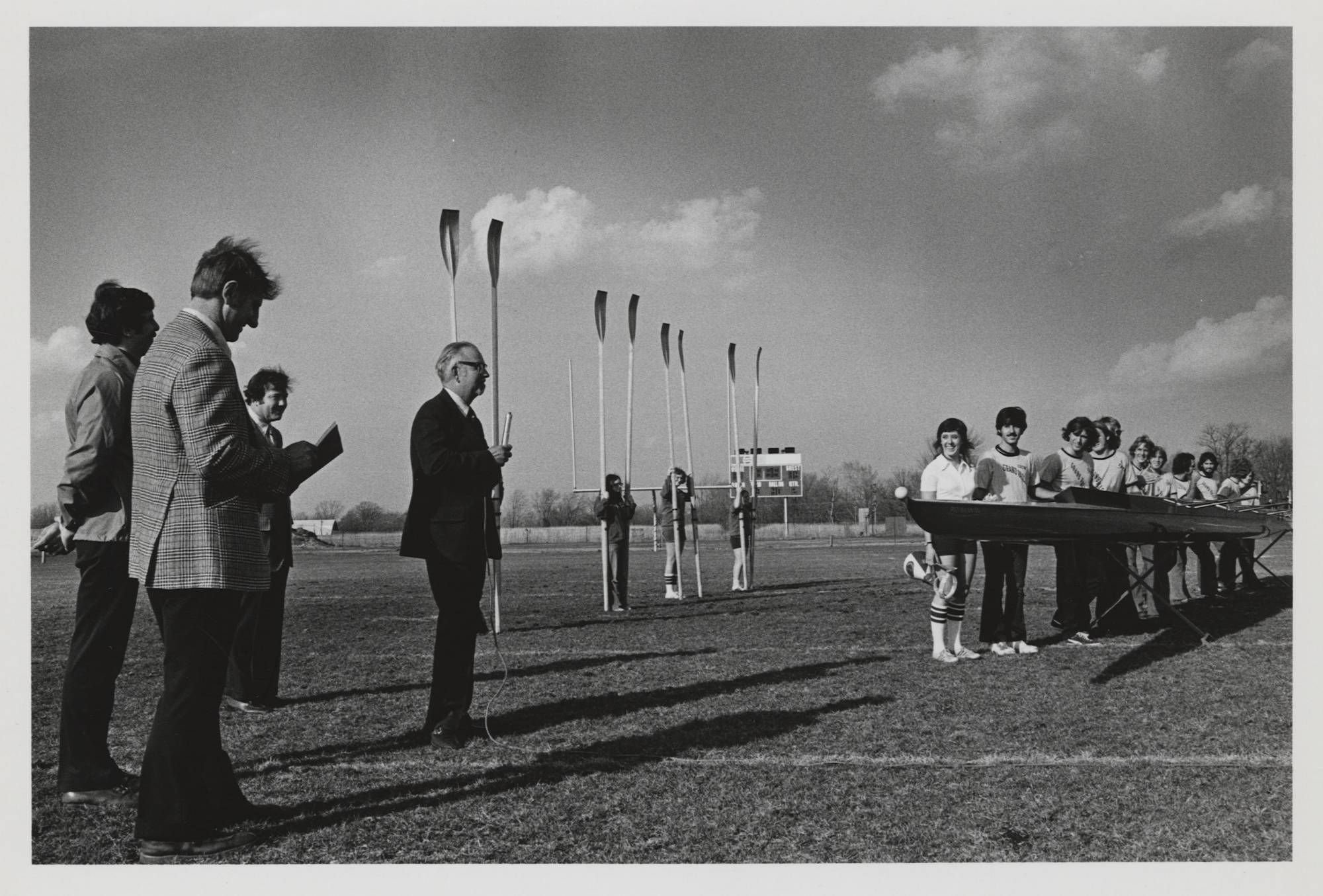 Members of the rowing team stand on the football field with university officials to dedicate a new rowing shell